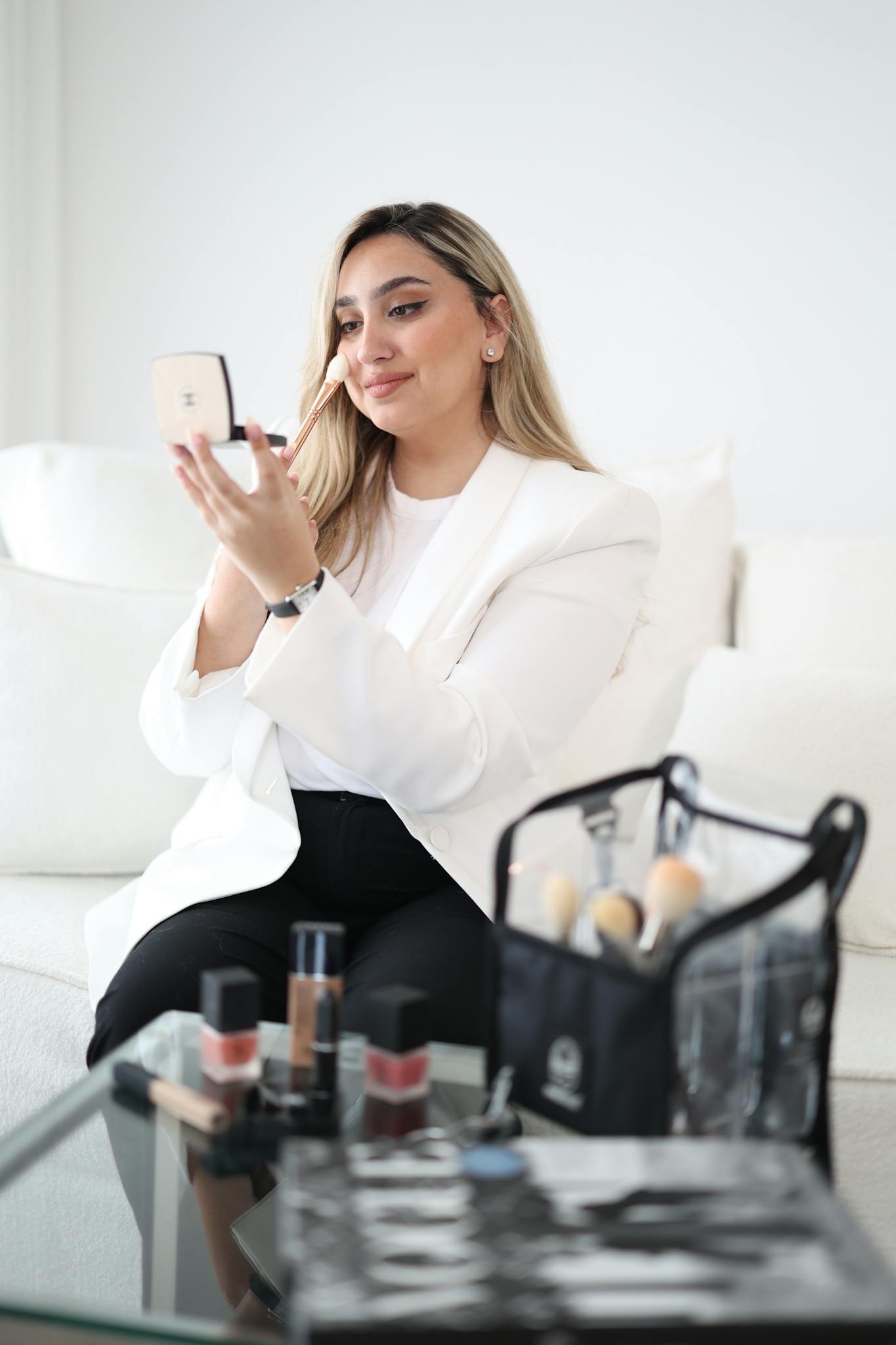 Woman seated applying makeup with a brush and mirror, showcasing cosmetics on a table.